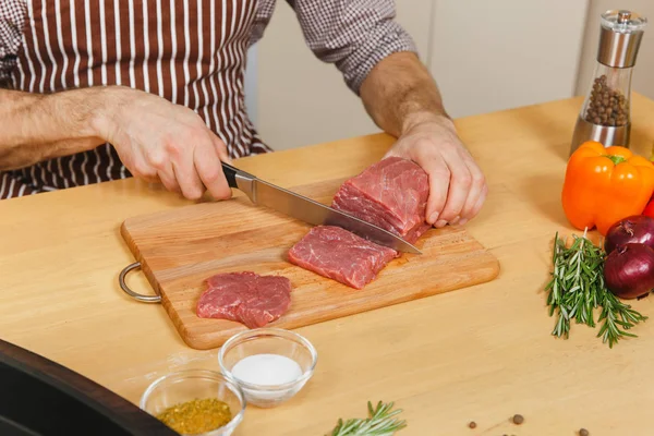 Close up jovem caucasiano em avental sentado à mesa com legumes, cozinhando em casa preparando estaca de carne de porco, carne bovina ou cordeiro, em cozinha leve com superfície de madeira, cheia de utensílios de cozinha chiques . — Fotografia de Stock