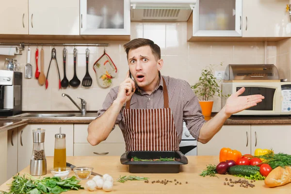 Perturbed stress young man in apron sitting at table with vegetables, talking on mobile phone, cooking at home preparing meat stake from pork, beef or lamb, in light kitchen with wooden surface. — Stock Photo, Image