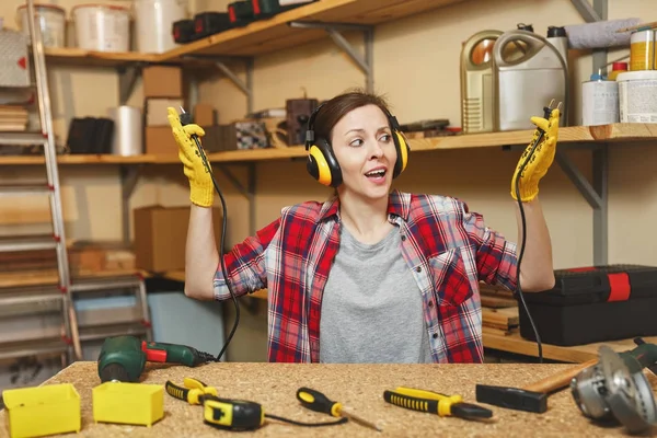 Beautiful crazy fun caucasian young woman in plaid shirt, noise insulated headphones working in carpentry workshop at table place, with two wires from electrical appliances like power drill and saw. — Stock Photo, Image