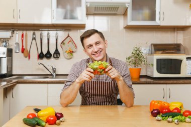 Gülümseyen Avrupa yakışıklı genç hafif modern mutfak kase sebze salatası ile masada oturan önlük, kahverengi gömlekli. Diyet kavramı. Sağlıklı bir yaşam tarzı. Evde yemek. Yemek hazırlamak.