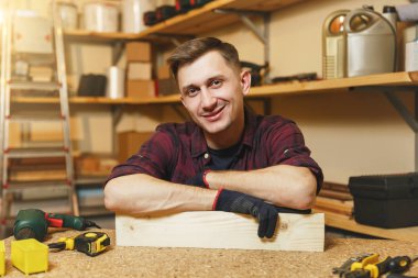 Yakışıklı beyaz genç adam ekose gömlek, siyah T-shirt, gülümseyen ahşap masa doğrama atölyesinde çalışan eldiven ile ahşap, farklı araçlar parça yerleştirin. Kopya alanı reklam için.