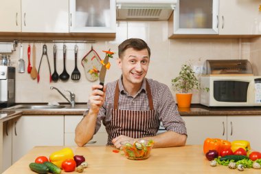 Işık mutfakta yemek çekici gülümseyen Beyaz delikanlı kase sebze salatası ile masada oturan önlük, kahverengi gömlekli. Diyet kavramı. Sağlıklı bir yaşam tarzı. Evde yemek. Yemek hazırlamak.