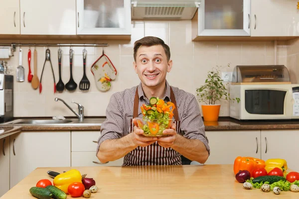 Schöner lustiger kaukasischer junger Mann in Schürze, braunes Hemd, der am Tisch sitzt und Gemüsesalat in einer Schüssel in der leichten modernen Küche aufwirft. Diätkonzept. Gesunder Lebensstil. Kochen zu Hause. Essen zubereiten. — Stockfoto