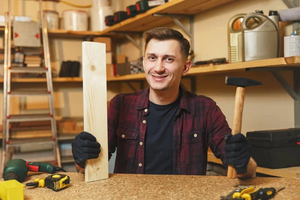 Bonito sorriso caucasiano jovem trabalhador homem em camisa xadrez, t-shirt preta, luvas de trabalho em oficina de carpintaria em lugar de mesa de madeira com pedaço de madeira, martelo, diferentes ferramentas . — Fotografia de Stock