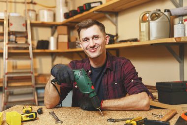 Yakışıklı beyaz genç adam ekose gömlek, siyah T-shirt, gülümseyen ahşap masa doğrama atölyesinde çalışan eldiven ile parça matkap, farklı araçlar yer.