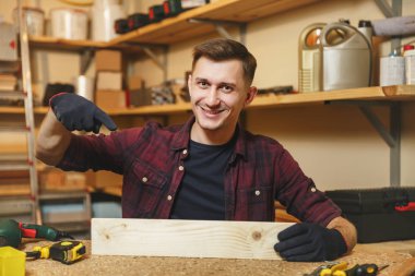 Yakışıklı beyaz genç adam ekose gömlek, siyah T-shirt, gülümseyen ahşap masa doğrama atölyesinde çalışan eldiven ile ahşap, farklı araçlar parça yerleştirin. Kopya alanı reklam için.