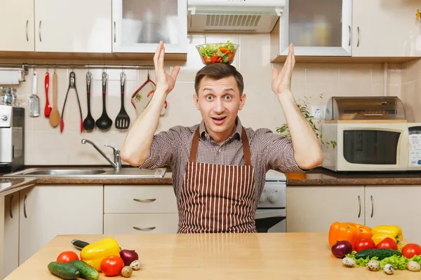 Attraktive lustige kaukasische junge Mann in Schürze, braunes Hemd sitzt am Tisch mit Gemüsesalat in Schüssel auf Kopf in heller Küche. Diätkonzept. Gesunder Lebensstil. Kochen zu Hause. Essen zubereiten. — Stockfoto