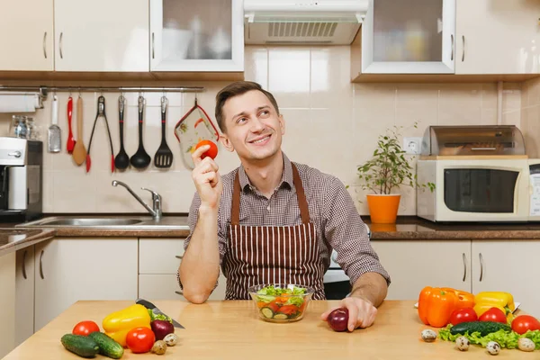 Jovem caucasiano divertido em avental, camisa marrom sentada à mesa, escolhendo entre cebola e tomate, cortando salada de legumes com faca na cozinha leve. Conceito de dieta. Cozinhar em casa. Preparar alimentos . — Fotografia de Stock