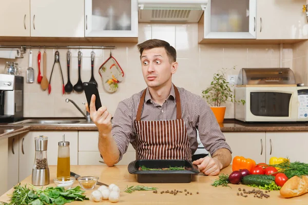 Perturbed stress young man in apron sitting at table with vegetables, talking on mobile phone, cooking at home preparing meat stake from pork, beef or lamb, in light kitchen with wooden surface. — Stock Photo, Image