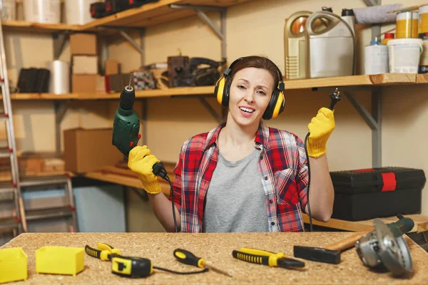 Jeune femme perplexe en chemise à carreaux gris T-shirt bruit casque isolé gants jaunes travaillant dans l'atelier de menuiserie à la place de la table en bois avec morceau de bois, différents outils, perceuse électrique, fil . — Photo