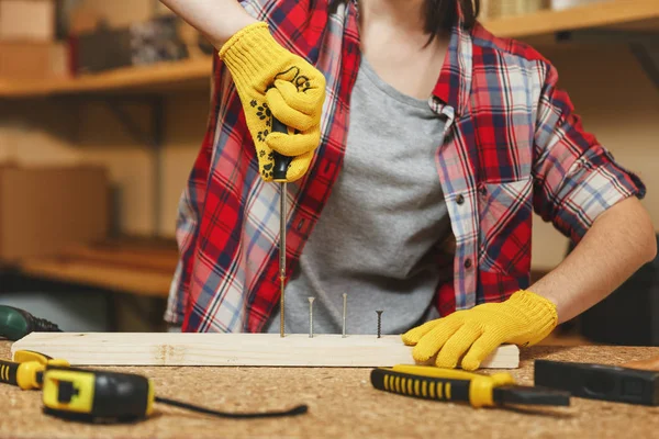 Gros plan de jeune femme caucasienne en chemise à carreaux, T-shirt gris, gants jaunes torsadés par vis tournevis, travail en atelier de menuiserie à la place de la table en bois avec morceau de bois, différents outils . — Photo