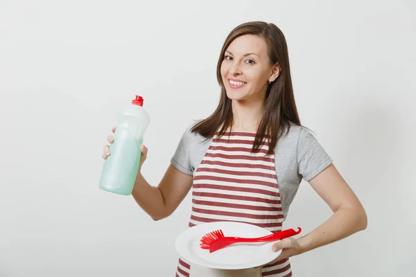 Casalinga in grembiule rigato isolata su sfondo bianco. Donna governante tiene liquido pulitore bottiglia, spazzola rossa per lavare i piatti, piatto rotondo vuoto bianco. Bottiglia con spazio per la copia pubblicitaria . — Foto Stock