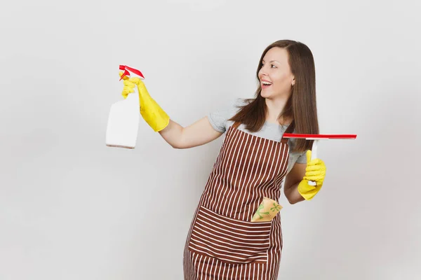 Young smiling housewife in yellow gloves, striped apron, cleaning rag in pocket isolated on white background. Housekeeper woman holding squeegee, spray bottle with cleaner liquid. Bottle copy space. — Stock Photo, Image