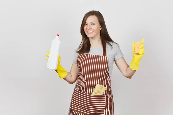 Junge Hausfrau in gelben Handschuhen, gestreifter Schürze, Putzlappen in der Tasche isoliert auf weißem Hintergrund. Frau spreizt Hände, hält Flasche mit Reinigungsflüssigkeit, Schwamm. Kopierfläche für Werbung. — Stockfoto