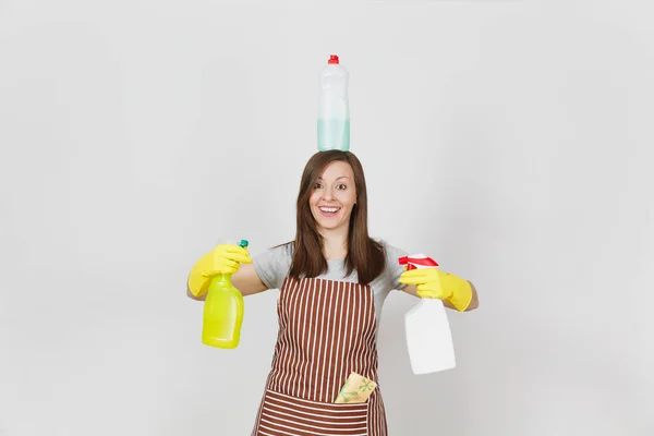 Fun housewife in yellow gloves, striped apron, cleaning rag in pocket on white background. Woman holding in hands, on head bottles with cleaner liquid for washing dishes. Copy space for advertisement. — Stock Photo, Image
