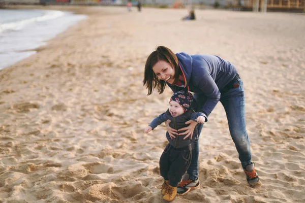 Jovem mulher jogando no fundo da praia de areia do mar com pouco menino bonito criança, bom humor. Mãe, filho pequeno no dia ensolarado. Paternidade, férias em família, 15 de maio, amor, pais, conceito de crianças — Fotografia de Stock