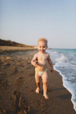 Küçük şirin çocuk bebek çocuk pantolon içinde yürümek ve denizde kum yaz sahilde, dağ arka plan çalıştırmak mutlu eğlence gülümseyen. Küçük çocuk oğlu açık havada oynamak. Aile günü 15 Mayıs, sevgi, anne, çocuk kavramı