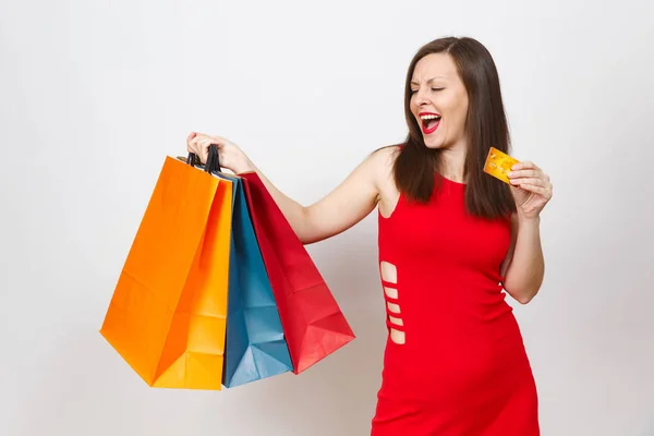 Mulher de cabelo castanho jovem bastante impressionante na moda em vestido vermelho segurando cartão de crédito, pacotes multicoloridos com compras depois de fazer compras isoladas em fundo branco. Espaço de cópia para propaganda . — Fotografia de Stock