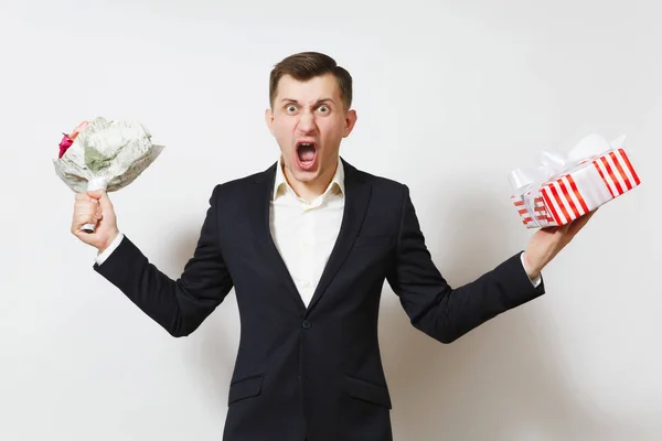 Young crazy angry perturbed man in suit holding bouquet of roses flowers, red present box with gift isolated on white background. St. Valentine's Day, International Women Day birthday holiday concept. — Stock Photo, Image
