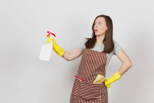 Young smiling housewife in yellow gloves, striped apron, cleaning rag, squeegee in pocket isolated on white background. Housekeeper woman holding spray bottle with cleaner liquid. Bottle copy space. — Stock Photo, Image