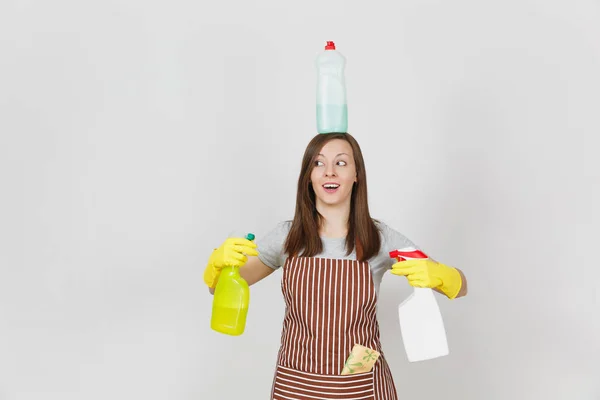 Fun housewife in yellow gloves, striped apron, cleaning rag in pocket on white background. Woman holding in hands, on head bottles with cleaner liquid for washing dishes. Copy space for advertisement. — Stock Photo, Image