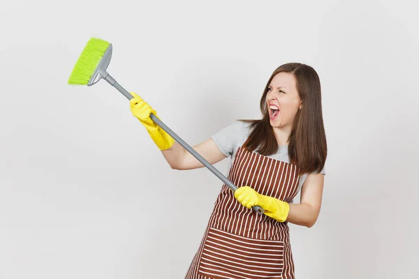 Young crazy housewife in striped apron, yellow gloves isolated on white background. Fun housekeeper woman cleaning maid holding and sweeping with broom. Copy space for advertisement. Advertising area. — Stock Photo, Image