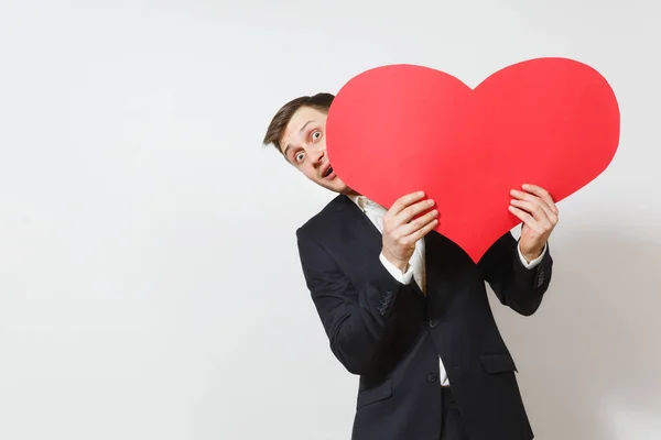 Joven hombre divertido en traje escondido detrás de un gran corazón rojo aislado sobre fondo blanco. Copiar espacio, publicidad. Lugar para el texto. Día de San Valentín, Día Internacional de la Mujer, cumpleaños, concepto de fiesta . —  Fotos de Stock