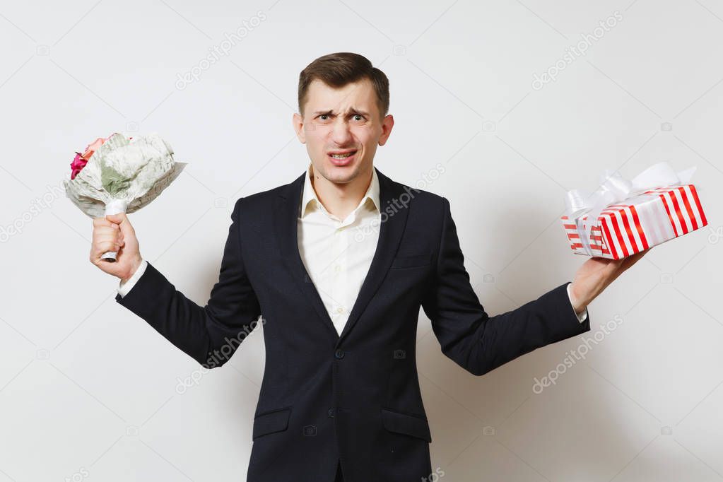 Young crazy angry perturbed man in suit holding bouquet of roses flowers, red present box with gift isolated on white background. St. Valentine's Day, International Women Day birthday holiday concept.