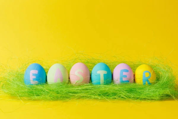 Fila de seis pastel monofônico colorido pintado ovos de Páscoa com inscrição Páscoa em grama verde isolado no fundo amarelo. Espaço de cópia para propaganda. Com lugar para SMS. Conceito de férias . — Fotografia de Stock