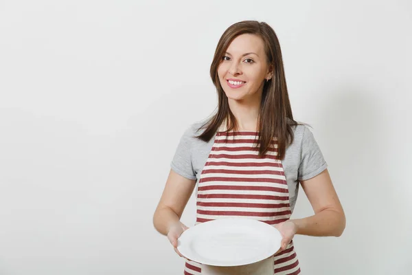 Jovem atraente sorridente morena caucasiana dona de casa em avental listrado, cinza t-shirt isolada no fundo branco. Mulher bela governanta segurando prato branco vazio. Espaço de cópia para propaganda . — Fotografia de Stock