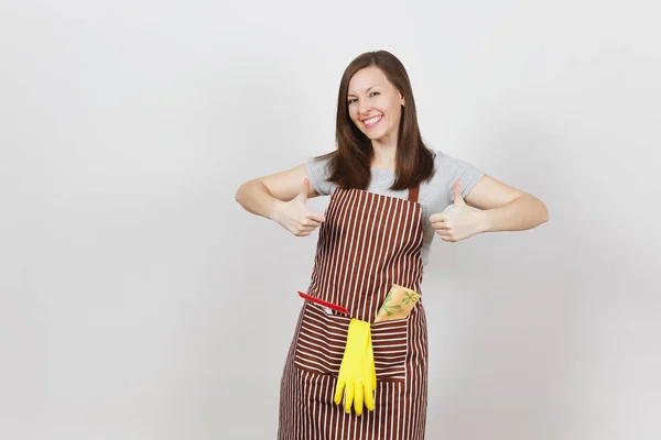 Jeune femme au foyer souriante en tablier rayé avec chiffon nettoyant, raclette, gants jaunes dans une poche isolée sur fond blanc. Jolie femme de ménage montrant pouces vers le haut regardant caméra. Pour la publicité — Photo