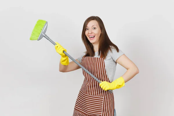 年轻微笑的家庭主妇在条纹围裙, 黄色手套隔离在白色背景。有趣的管家女清洁工拿着扫帚扫。广告广告区复制空间 — 图库照片
