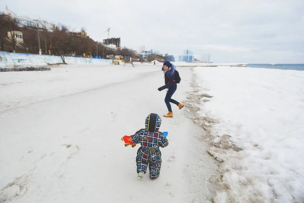 Donna giocare a mare neve spiaggia sfondo con piccolo bambino carino bambino in tuta con auto giocattolo, pala. Mamma, figlio bambino divertente il giorno d'inverno. Vacanza in famiglia, 15 maggio, amore, genitori, concetto bambini . — Foto Stock