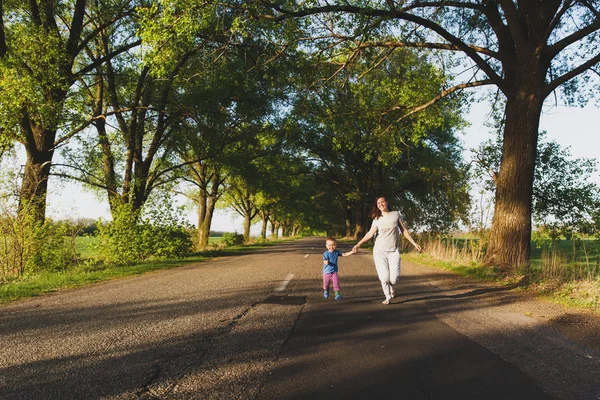 Young woman, little cute child boy walk on road with trees on the roadside, catch up, run in warm summer day. Mother, kid son rest. Parenthood, family day, 15 of may, love, parents, children concept. — Stock Photo, Image