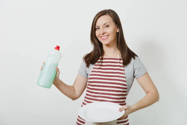 年轻的家庭主妇在条纹围裙隔离白色背景。女管家拿着瓶子用干净的液体洗碗, 白色的空圆盘子。广告复制空间瓶. — 图库照片