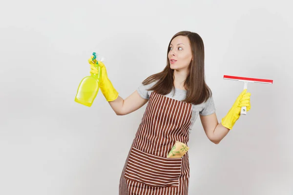 Young smiling housewife in yellow gloves, striped apron, cleaning rag in pocket isolated on white background. Housekeeper woman holding squeegee, spray bottle with cleaner liquid. Bottle copy space. — Stock Photo, Image