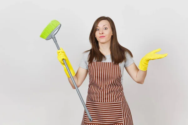 Young perplexed housewife in striped apron, yellow gloves isolated on white background. Fun housekeeper woman cleaning maid holding and sweeping with broom. Copy space advertisement. Advertising area. — Stock Photo, Image