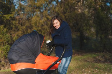 Genç kadın mavi ceket sonbahar doğa, yeşil ağaçlar üzerinde küçük çocuk bebek turuncu bebek arabası, çay veya kahve ile sonbahar Park'ta yürüyordunuz. Anne ve çocuk. Ebeveynlik, aşk, Aile, çocuk kavramı.