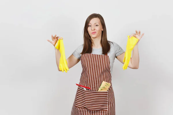 Young sad upset tired shocked housewife in striped apron with cleaning rag in pocket isolated on white background. Housekeeper woman holds smell yellow gloves in spreading hands. For advertisement.