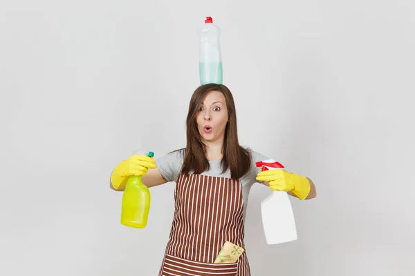 Fun housewife in yellow gloves, striped apron, cleaning rag in pocket on white background. Woman holding in hands, on head bottles with cleaner liquid for washing dishes. Copy space for advertisement. — Stock Photo, Image