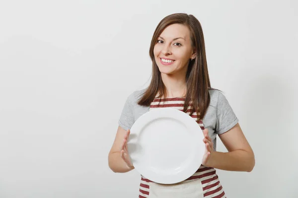 Joven atractiva morena sonriente ama de casa caucásica en delantal rayado, camiseta gris aislada sobre fondo blanco. Hermosa ama de llaves mujer sosteniendo plato blanco vacío. Copiar espacio para publicidad . —  Fotos de Stock