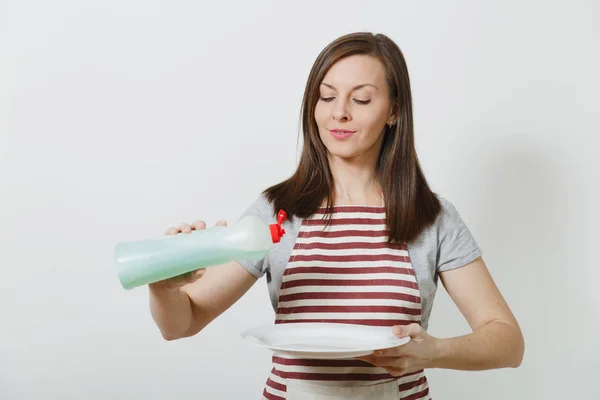 年轻的家庭主妇在条纹围裙隔离白色背景。女管家拿着瓶子用干净的液体洗碗, 白色的空圆盘子。广告复制空间瓶. — 图库照片