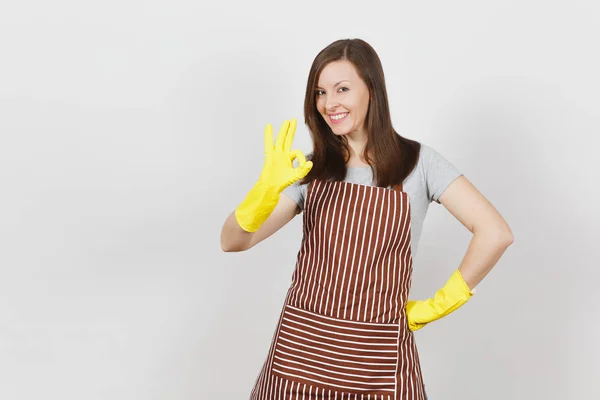 Jonge aantrekkelijke lachende brunette huisvrouw in gestreepte schort, gele handschoenen geïsoleerd op een witte achtergrond. Mooie huishoudster vrouw weergegeven: Ok gebaar op zoek camera. Ruimte voor reclame kopiëren. — Stockfoto