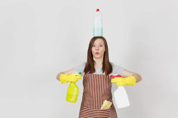 Divertente casalinga in guanti gialli, grembiule a righe, straccio detergente in tasca su sfondo bianco. Donna che tiene in mano, su bottiglie principali con liquido più pulito per lavare piatti. Copia spazio per pubblicità . — Foto Stock