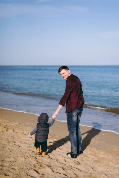 Jovem de pé no fundo da praia de areia do mar segurando mão pequeno menino bonito criança, olhando no horizonte. Pai, rapazinho. Dia de família 15 de maio, amor, pais, conceito de crianças. Visão traseira . — Fotografia de Stock