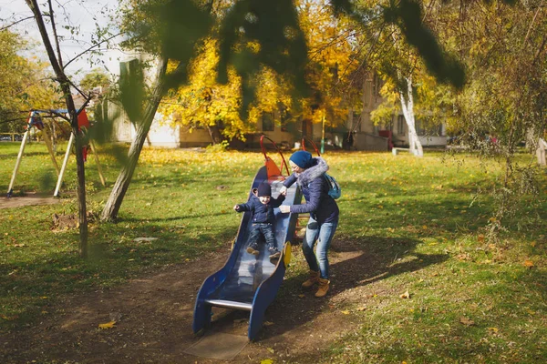 Mujer joven caminar en verde amarillo otoño otoño parque con pequeño niño lindo bebé en la naturaleza. Madre, hijo hijo descanso divertido, montar diapositivas de los niños en el patio de recreo. Día de la familia 15 de mayo, amor concepto de los padres . — Foto de Stock