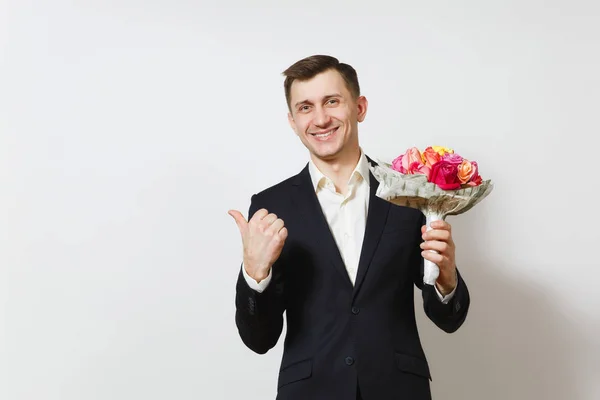 Молодой улыбающийся мужчина в костюме, указывая пальцем в сторону, держит букет красивых цветов роз, изолированных на белом фоне. День святого Валентина, Международный женский день, день рождения, праздничная концепция — стоковое фото