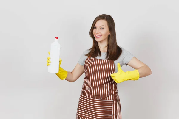 Joven ama de casa sonriente con guantes amarillos, delantal a rayas aislado sobre fondo blanco. Mujer mostrando el pulgar hacia arriba, sosteniendo la botella con líquido limpiador para lavar y limpiar. Copiar espacio para publicidad . —  Fotos de Stock