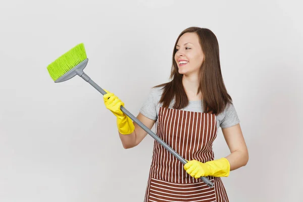 Unga leende hemmafru i randigt förkläde, gula handskar isolerad på vit bakgrund. Kul hushållerska kvinna rengöring maid holding och svepande med kvast. Kopiera utrymme för reklam reklam område — Stockfoto