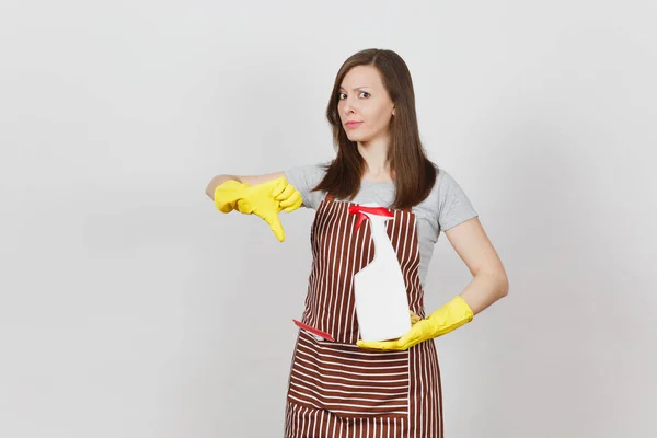 Joven triste triste ama de casa cansada en guantes amarillos delantal rayado limpiando rag squeegee en bolsillo aislado sobre fondo blanco. Mujer muestra pulgar hacia abajo sostiene la botella de aerosol con líquido más limpio. Copiar espacio . —  Fotos de Stock